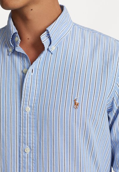Lyseblå og hvit stripete skjorte med knapper og krage, samt en liten brodert logo på brystet. Laget av lett stoff.