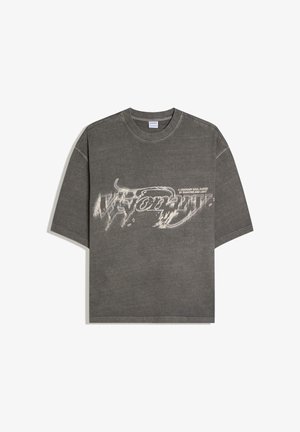 Sötétszürke, túlméretezett póló, torzított fehér szöveggel "Visionary" és kisebb szöveggel "A visionary soul guided by shadows and light" elöl.