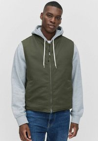 Olivgrön ärmlös jacka med en grå luvtröja under, med dragkedja framtill och snoddar, kombinerad med blå jeans.