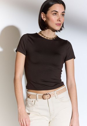 Donna dai capelli corti e scuri che indossa una t-shirt nera aderente, pantaloni beige con cintura beige e una collana d'oro spessa, guarda a destra.