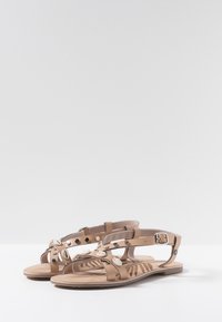 Sandalias planas color bronce con correas de tobillo con hebilla, decoradas con pequeñas conchas y tachuelas metálicas, con una plantilla estampada.