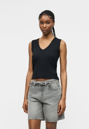 Jeune femme aux cheveux tressés portant un haut noir sans manches à col en V et un short en jean gris clair avec une ceinture nouée, debout devant un fond uni.