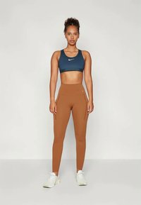Modrá športová podprsenka s bielym logom, skombinovaná s hnedými legínami s vysokým pásom a bočnými vreckami. Model nosí biele atletické topánky.