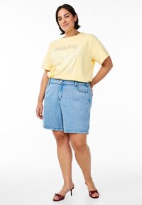 Camiseta amarilla con gráfico de mariposa, combinada con pantalones cortos de mezclilla azul claro. La modelo lleva sandalias de tacón rojas; un atuendo casual con un corte relajado.