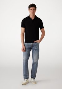 Camisa polo negra con cuello, combinada con pantalones vaqueros de mezclilla azul claro. El modelo lleva zapatillas blancas. Diseño simple y entallado con mangas cortas.