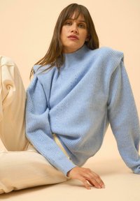 Vaaleansininen ribbikuvioinen neuletakki, jossa on korkea kaulus ja tiputettu olkalinja, yhdistettynä kermavärisiin leveälahkeisiin housuihin. Sujuva pinta, rento istuvuus.