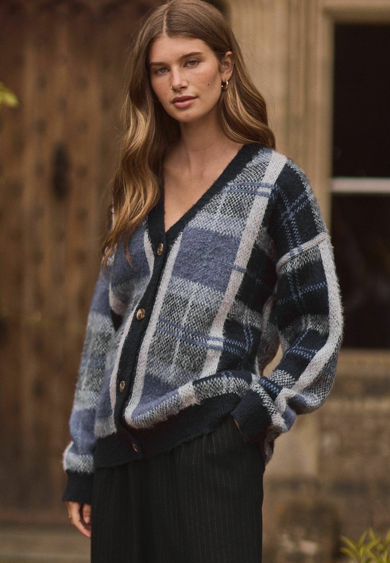Gilet tricoté avec un motif à carreaux bleu et gris, col en V, bord noir et boutons bruns. Matériau en laine douce et texturée.
