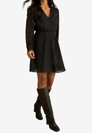 Kobieta ubrana w czarną, teksturowaną sukienkę do kolan z długimi rękawami i dekoltem w serek, w połączeniu z czarnymi, sięgającymi kolan, butami na obcasie.