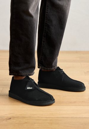 Person trägt schwarze Wildleder-Schnürschuhe und dunkelgraue Hose und steht auf einem hellen Holzboden.