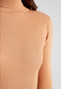 Pull à col roulé côtelé de couleur pêche, avec une coupe slim et un tissu texturé aux lignes verticales. Aucun motif ni ornement visible.