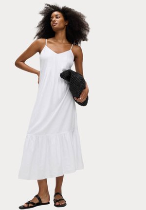 Vestido midi blanco sin mangas con un dobladillo en capas, hecho de una tela ligera. La modelo sostiene un bolso negro de mimbre. Las sandalias presentan un diseño entrecruzado.