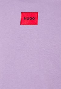 T-shirt di cotone lilla con una toppa rettangolare rossa testurizzata con la scritta "HUGO" in nero, posizionata sul petto.