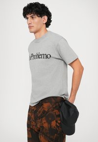 Graues Baumwoll-T-Shirt mit schwarzem Text, der "No Problemo" sagt. Kombiniert mit gemusterten braunen Hosen und einer schwarzen Baseballmütze.