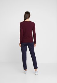 Femme de dos, portant un haut bordeaux à manches longues, un pantalon bleu marine et des baskets gris clair sur un fond blanc.