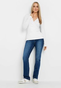 Weißes Langarm-Wickeloberteil, kombiniert mit blauen gerade geschnittenen Jeans und weißen Sneakers. Glattes Material, minimale Details und taillierte Form.