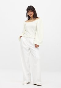 Maglietta bianca a maniche lunghe con texture e design off-the-shoulder, abbinata a pantaloni bianchi a vita alta, morbidi e scarpe flat a punta.