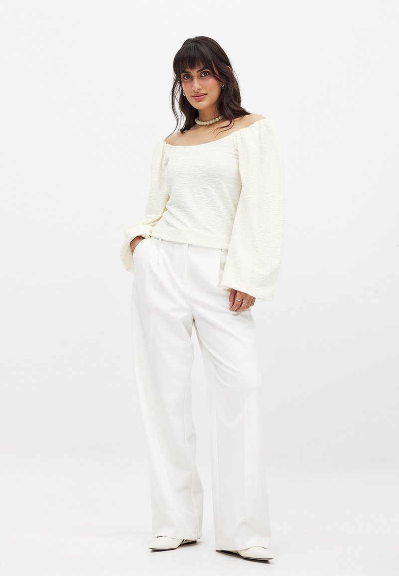 Maglietta bianca a maniche lunghe con texture e design off-the-shoulder, abbinata a pantaloni bianchi a vita alta, morbidi e scarpe flat a punta.