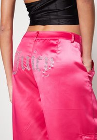 Rosa satinbyxor med en blanka yta, prydda med rhinstenar som stavar "Juicy" på bakfickan. Skräddarsydd design med sidofickor.