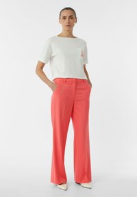 Biała bawełniana koszulka z małym koralowym wzorem kieszeni, w połączeniu z koralowymi spodniami o szerokich nogawkach. Modelka nosi beżowe buty z ostrym noskiem.