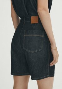 Shorts en denim foncé avec une taille haute, des coutures visibles de couleur contrastante et une étiquette en cuir à l'arrière de la ceinture.