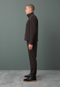 Giacca zip-up marrone scuro con colletto alto, abbinata a pantaloni coordinati e sneakers nere. Il tessuto appare morbido con una finitura testurizzata.