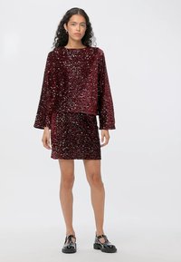 Bordeaux-kleurige pailletten crop top en bijpassende rokset. De stof heeft een glanzende textuur. De mouwen zijn lang en de rok is aansluitend. Zwarte schoenen.