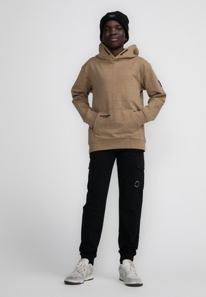 Tan hoodie met een gestructureerde logo-opdruk, gecombineerd met zwarte cargo broeken met zakken, en witte sneakers met grijze accenten.
