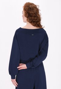Maglietta a maniche lunghe blu navy con scollo ampio, maniche plissettate e dettagli di branding discreti sulla schiena. Tessuto morbido e leggero.
