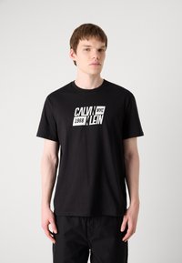 Fekete pamut póló, kerek nyakkivágással, elöl fehér grafikával, amelyen a "CALVIN KLEIN NYC 1968" felirat található. Rövid ujjak és laza fazon jellemzi.