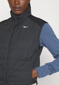 Gilet rembourré noir avec col montant, fermeture éclair et logo Nike blanc. Présente des épaules noires contrastées et une chemise à manches longues bleu marine en dessous.