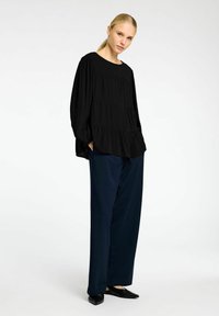 Sort langærmet bluse med rund halsudskæring og lagdelt design, parret med marineblå brede bukser og sorte fladsålede sko. Glat stofstruktur.