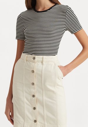 Vrouw draagt een zwart-wit gestreept T-shirt met korte mouwen, ingestopt in een crèmekleurige rok met knopen aan de voorkant en zakken.