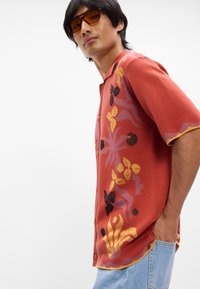 Rustfarget kortermet skjorte med knapper som har blomster- og sommerfugl-mønstre i rosa, svart og gul. Strukturert strikket stoff.