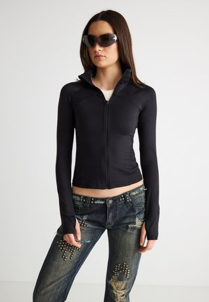 Kvinde iført sort tætsiddende lynlåstrøje, mørke revnede jeans med metalnitter og store sorte solbriller, stående mod en ensfarvet baggrund.