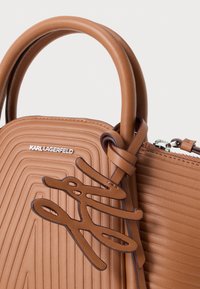 Brown leather handbag with embossed lines, a rounded handle, and a decorative logo accent. Features a top zipper closure and minimalist design.