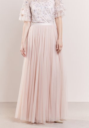 Femme porte une jupe maxi plissée rose clair avec un chemisier transparent orné de volants aux manches courtes.