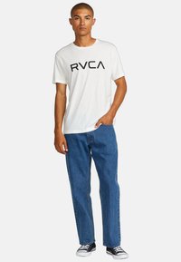 T-shirt en coton blanc avec le logo noir "RVCA", accompagnée de jeans en denim bleu. Le modèle porte des baskets noir et blanc.
