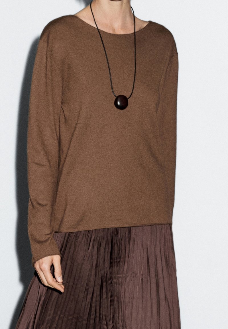 Personne portant un pull marron à manches longues, un collier pendentif rond foncé et une jupe plissée foncée sur un fond uni.