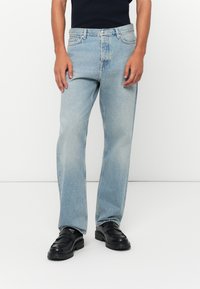 Man wearing light blue straight-leg jeans and black leather loafers standing against a white background.