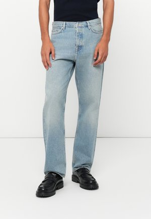 Un homme portant un jean droit bleu clair et des mocassins en cuir noir, se tenant devant un fond blanc.