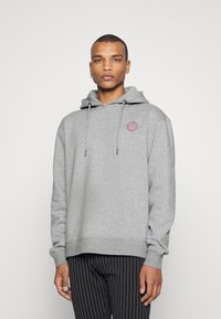 Grauer Hoodie mit einer Fronttasche, Kapuze mit Kordelzug und rotem sowie blauem Grafiklaster auf der Brust. Der Stoff wirkt weich und hat eine lockere Passform.