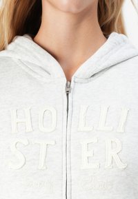 Lähedalt vaade isikule, kes kannab heledat halli küllatõmmatud kapuutsi, mille ees on valge õmmeldud "Hollister Surf Club" kiri.