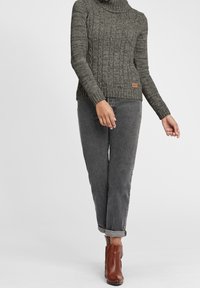 Grober, grauer Strickpullover mit hohem Kragen, gerippter Struktur und seitlichem Logo-Patch, kombiniert mit grauen, zulaufenden Hosen und braunen Stiefeletten.