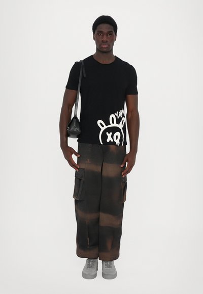 T-shirt noir avec un graphisme blanc, pantalon cargo marron ample avec poches, baskets grises et un petit sac à bandoulière noir. Fond plat et neutre.