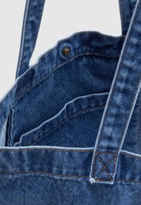 Μπλε τσάντα tote από τζιν σε κοντινό πλάνο που δείχνει τα ραμμένα χερούλια, την εσωτερική τσέπη και το μεταλλικό κουμπί πόρπης σε απλό φόντο.