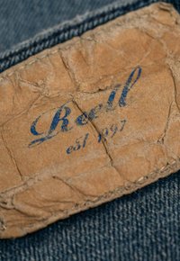 Etiqueta de cuero marrón desgastada con el texto azul "Reell est. 1997" cosido sobre tela de mezclilla azul.