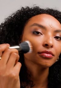 Femme aux cheveux bouclés appliquant du fond de teint sur sa joue avec un pinceau de maquillage, du fond de teint visible sur son visage et son cou.