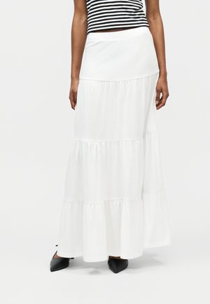 Femme portant une jupe maxi blanche à étages, des chaussures noires à bouts pointus, et un haut sans manches rayé noir et blanc, debout sur un fond blanc.