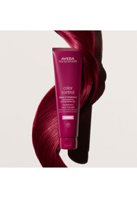 Traitement sans rinçage Aveda Color Control dans un tube fuchsia, avec du texte blanc, accompagné d'une mèche de cheveux magenta brillante à côté.