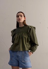 Blouse en coton vert texturé avec des épaules froncées et un motif œillet ; associée à un short en denim bleu clair avec des clous en métal.